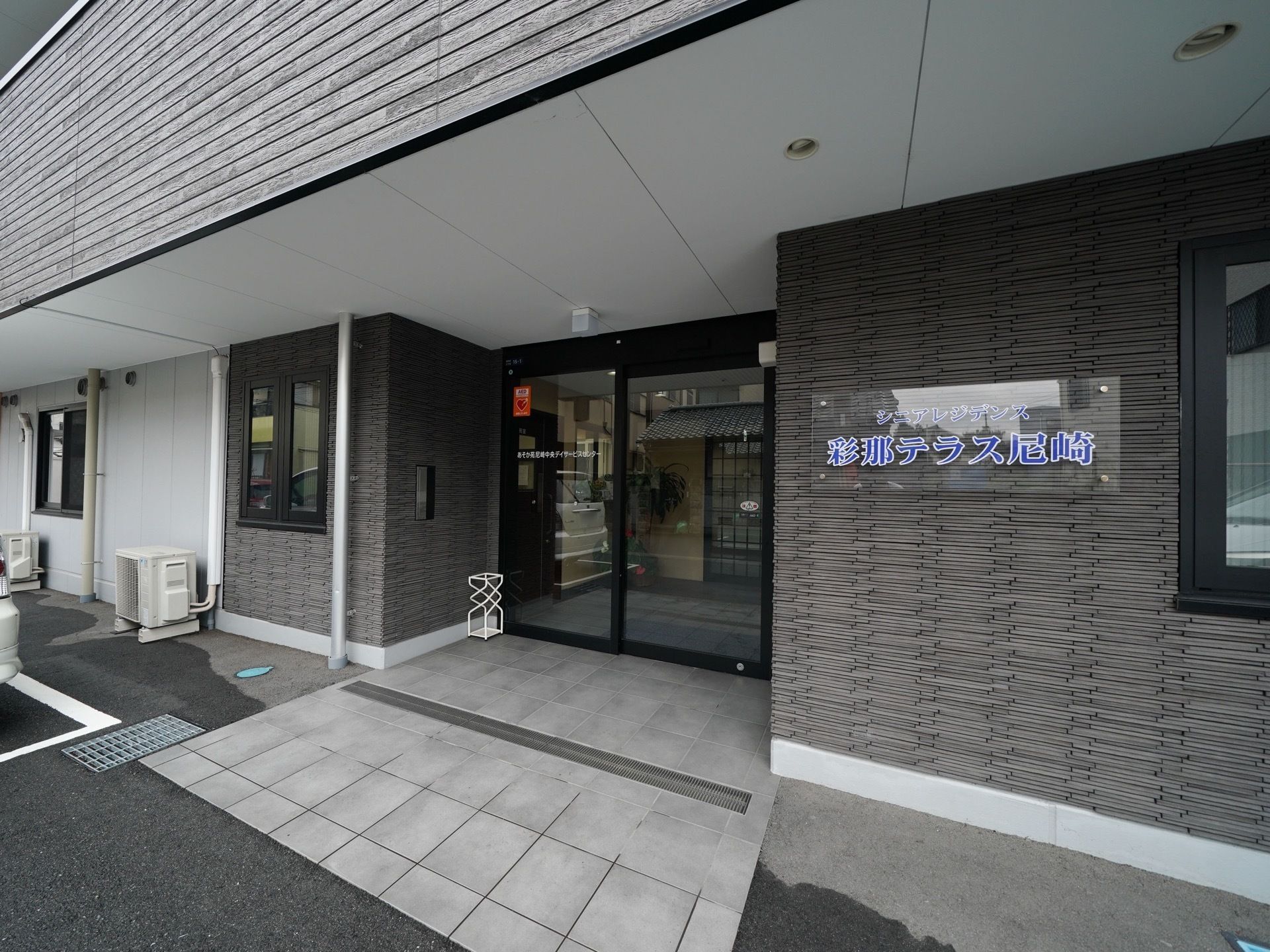玄関前のスペースには屋根を設置している。脇の壁には施設名を表示しており、透明のプレートに青色の文字で書かれている。