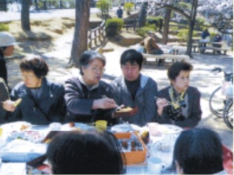 屋外でお弁当を広げた白いテーブルを囲んで会食している複数の人物