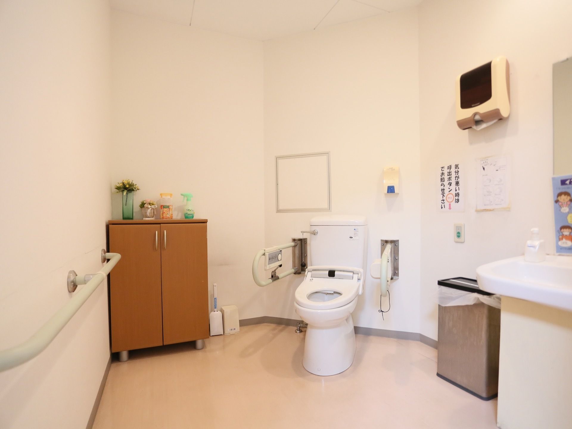 トイレの便座の周りには広々としたスペースがある。壁には長い手すりがあり、洗面台も近くに設置されている。