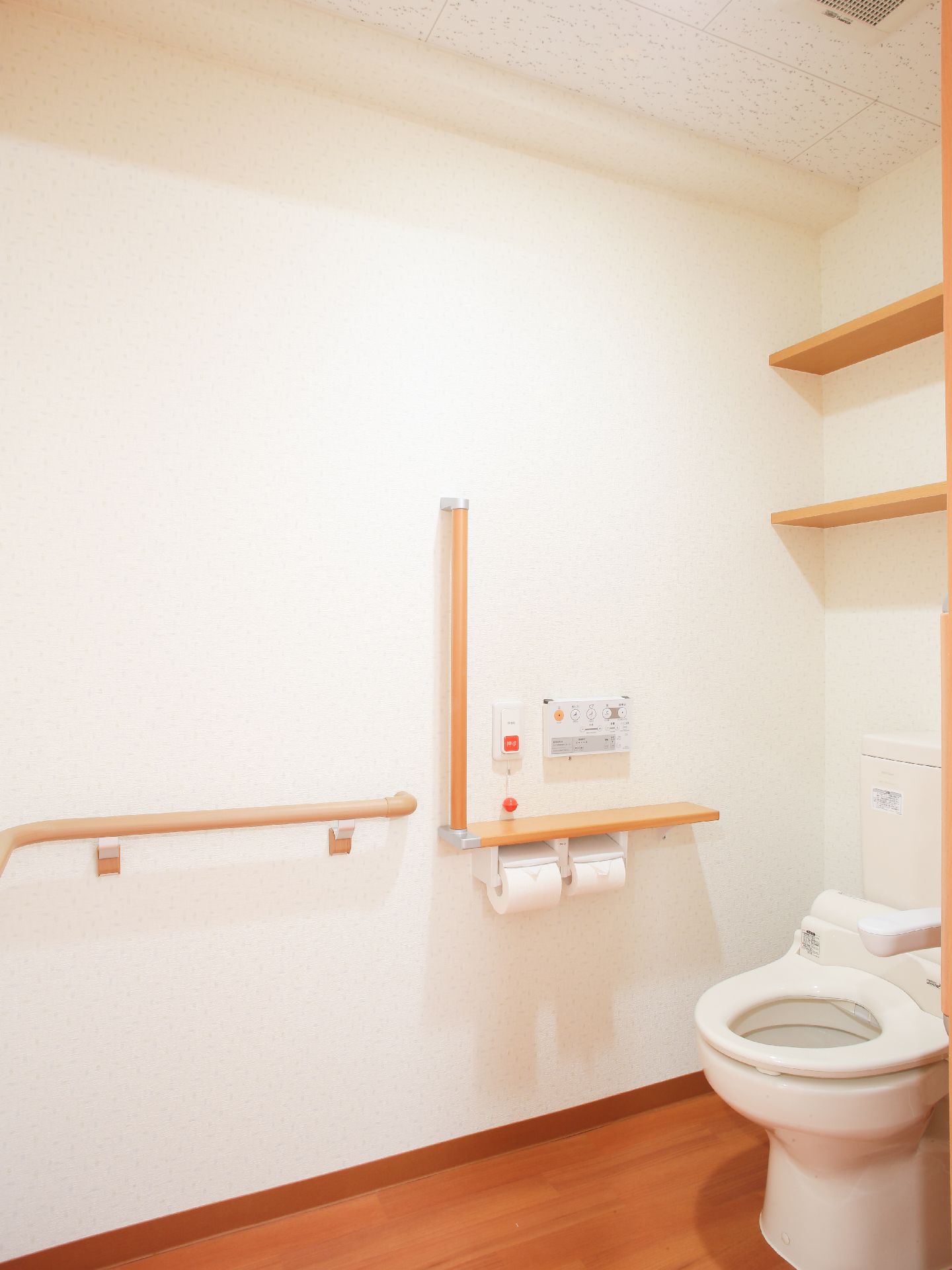白い壁に茶色のフローリングのトイレで、手すりが横についている。トイレットペーパーホルダーの上に棚がある。緊急時ブザーが壁に取り付けてある。