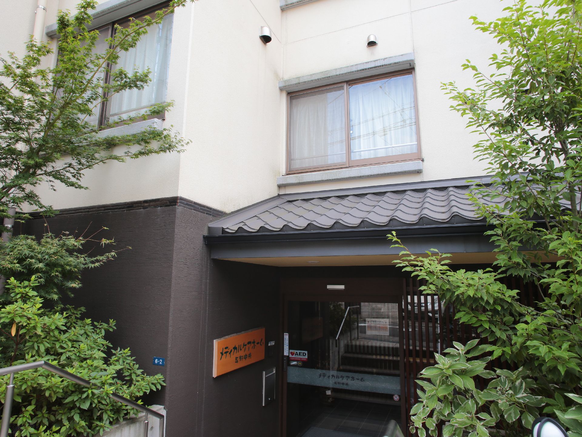 施設の玄関前で左右ともに植栽がしてある。玄関の左手に壁に呼び鈴が取り付けてある。瓦風の屋根が取り付けてある。
