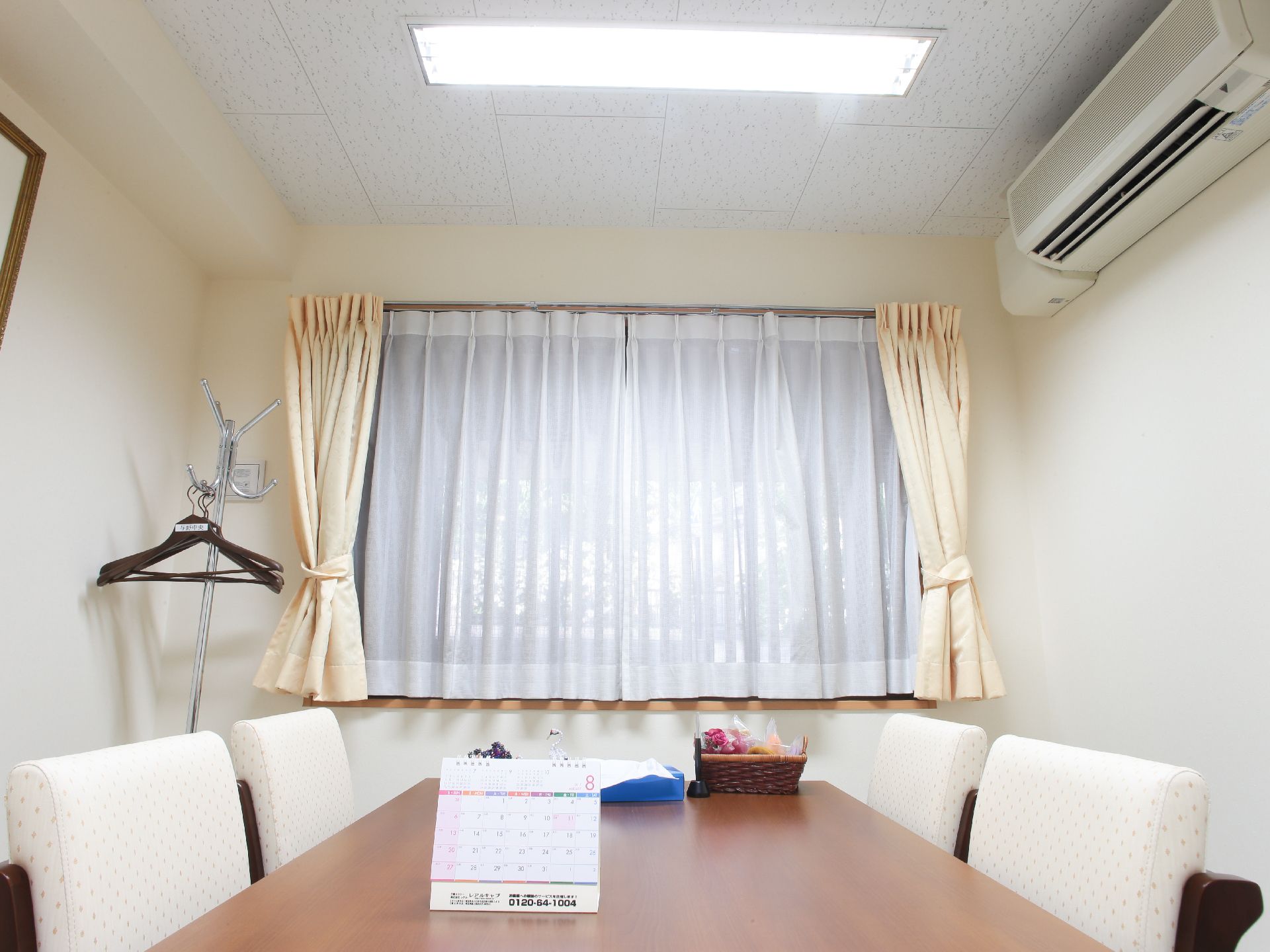 腰高窓がある部屋で、ハンガーポールが左手にある。レースと布のカーテンが取り付けてある。白い背もたれの椅子が四脚ある。