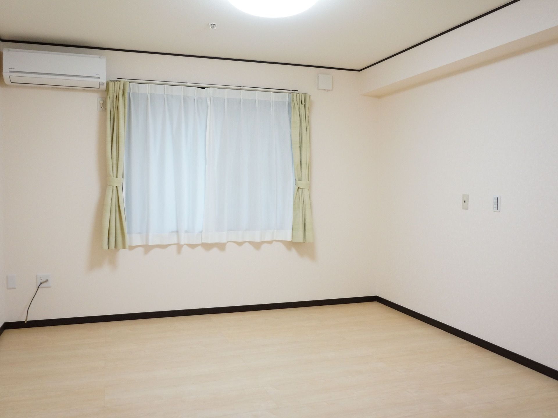居室にはレースと黄緑色のカーテンが掛かっている。薄い色のフローリングの床で白い壁にはエアコンが１台取り付けられている。