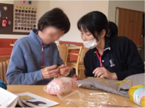 テーブルについて編み物をしている女性と、その手元をのぞき込んでいる女性スタッフ