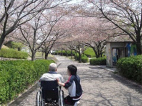 奥に桜並木、手前を植え込みに囲まれた通路で、車椅子を利用している人に屈んで話しかけている人物