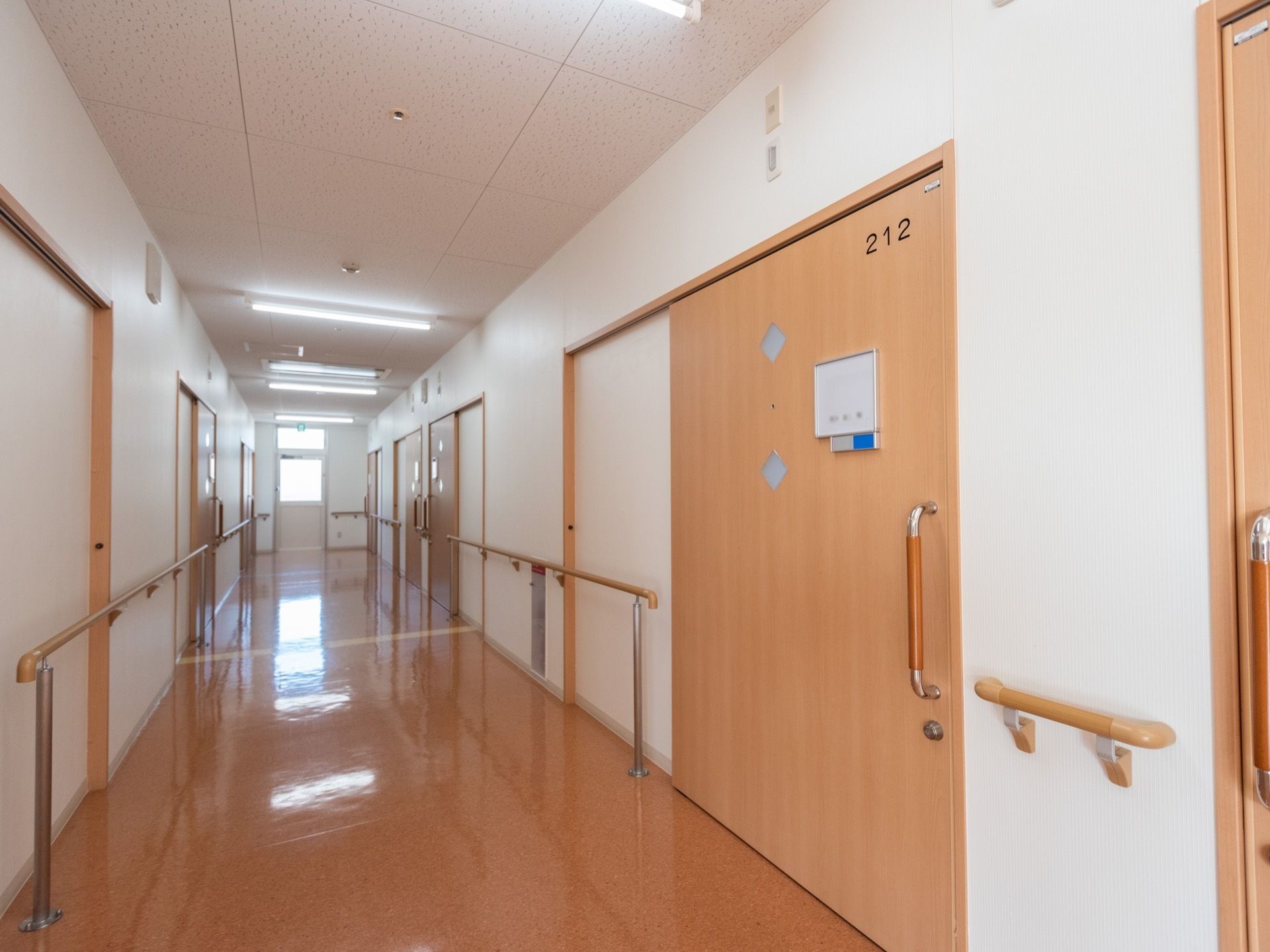 ゆったりとした施設の廊下である。廊下の壁には手すりが備え付けられ、幅のある居室の引き戸が設置されている。