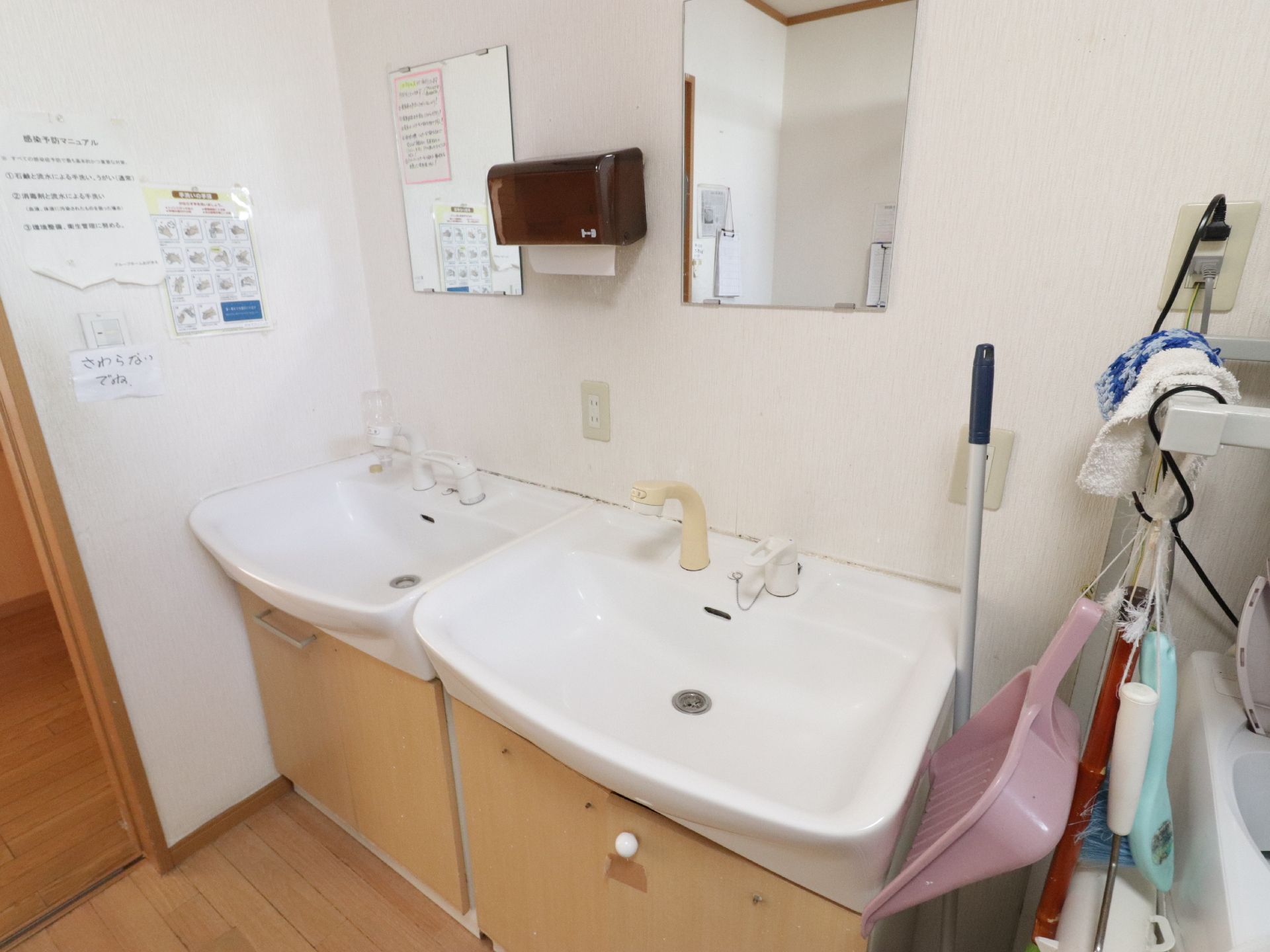 洗面台が2台並んでいる。前方には鏡が設置されている。左壁側には感染予防マニュアルや手洗いの手順が書かれた紙が貼られている。