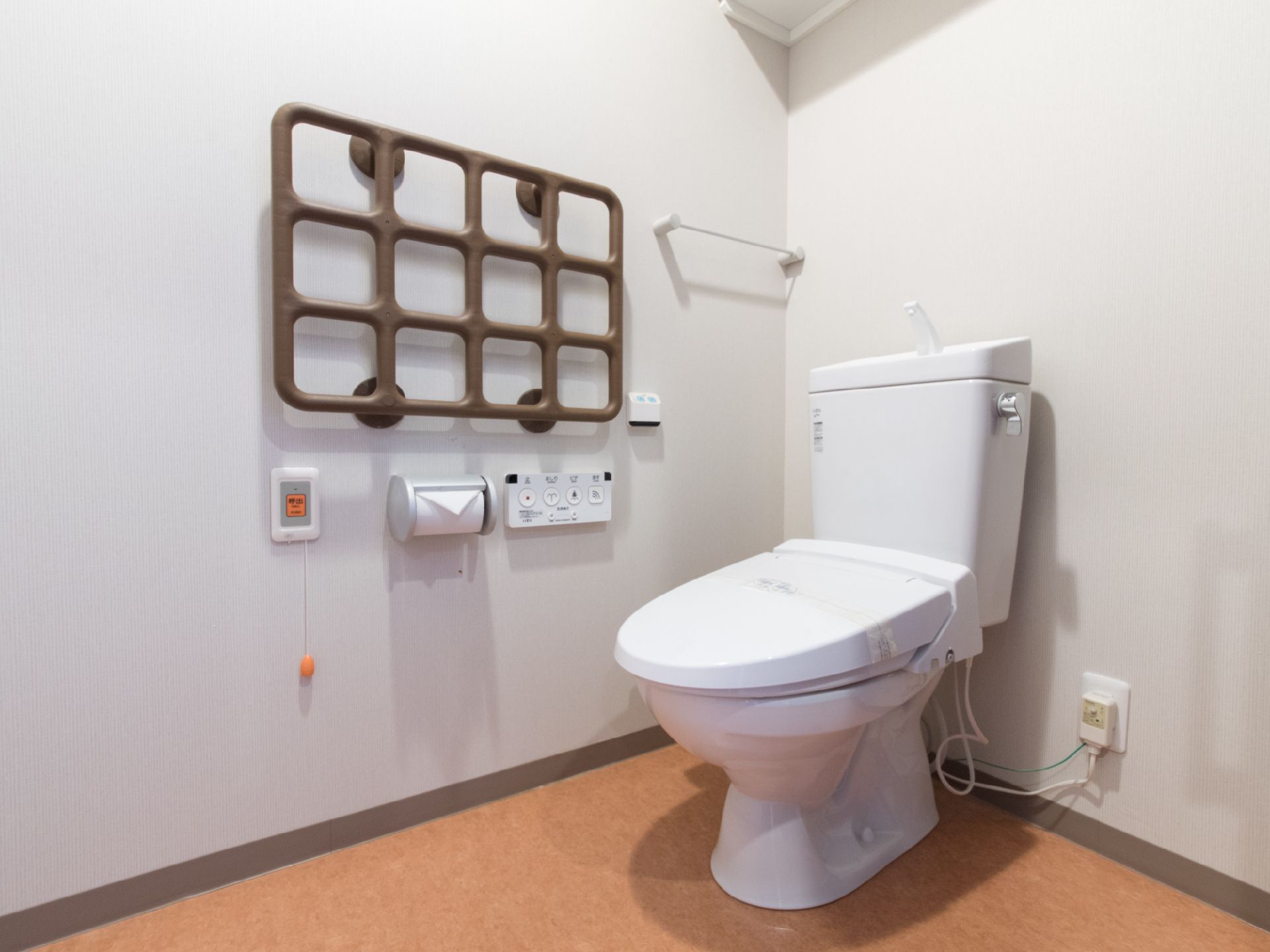オレンジ系の床にトイレが設置されている。右側の壁には格子状の手すりがあり、その下に操作パネルとナースコールがある。
