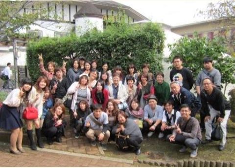 茶色い円錐状の屋根のある白い建物と植木の前で集合写真を撮るグループ