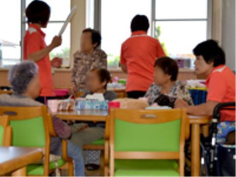 テーブルについている4人の高齢者と、それを介助しているオレンジのシャツを着た3人のスタッフ
