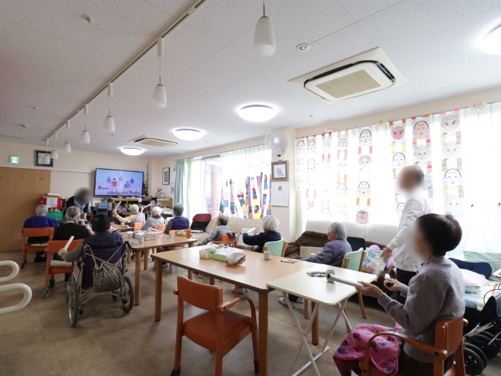 共有スペースには入居者様が集まって席に座っている。手には筒のようなものを持っている。前方にはテレビがある。