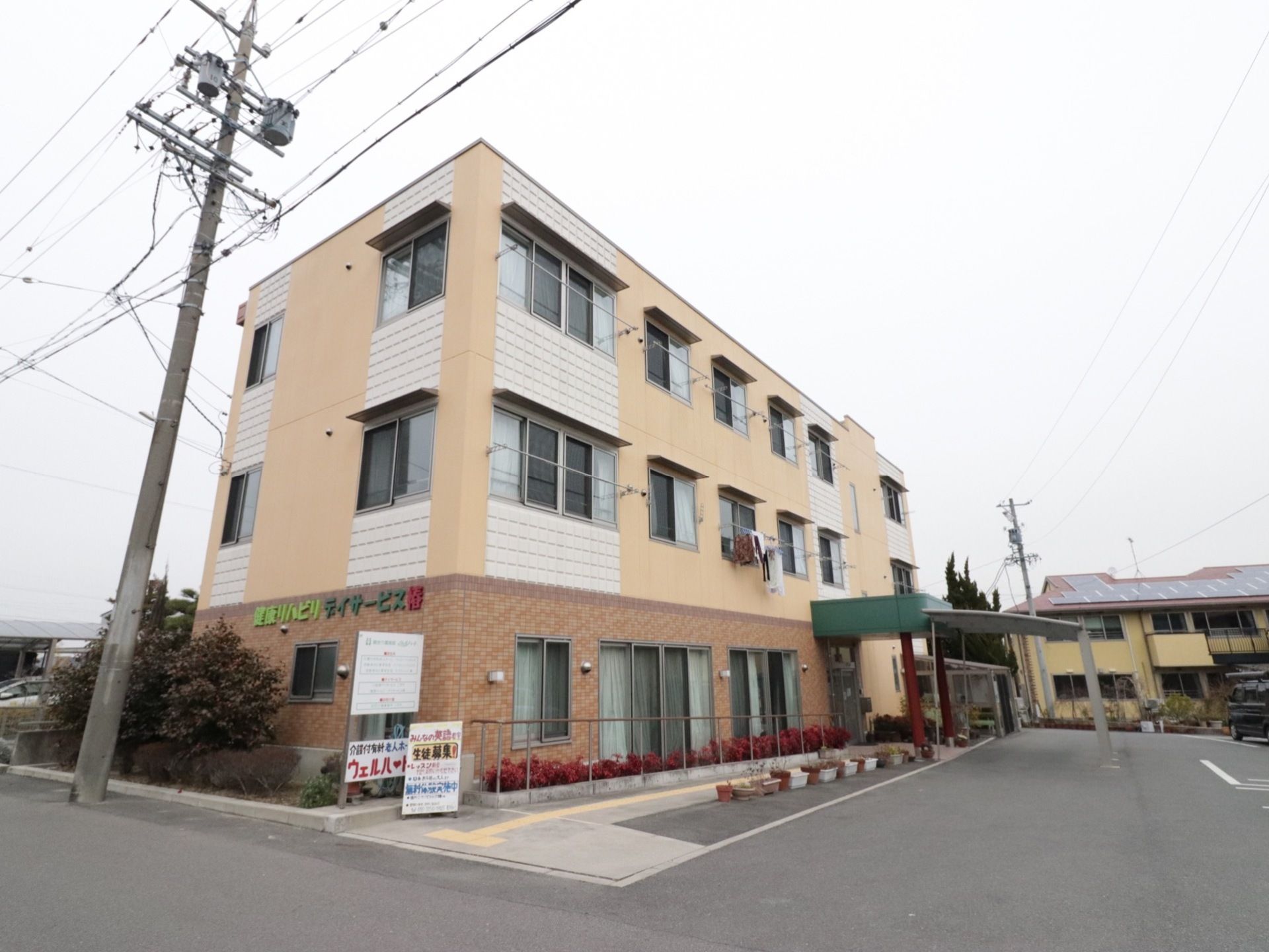 施設は三階建てである。施設建物の周りには花壇やプランターが並んでいる。入口には大きな屋根が取付けられている。