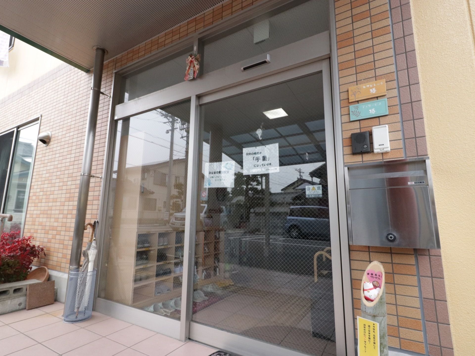 玄関入口にはガラス扉が設置されていて、施設内が見える。扉の右側にはインターフォン、ポスト、左側には傘立てが置かれている。