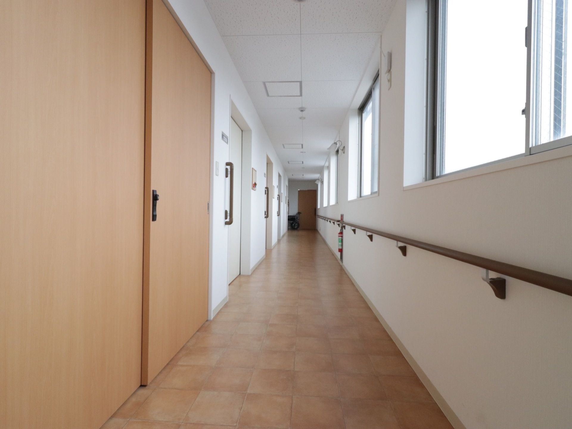 通路の右側には窓と手すりがあり、真っ直ぐに続いている。消火器もある。左側には茶色と白色の部屋の扉が並んでいる。