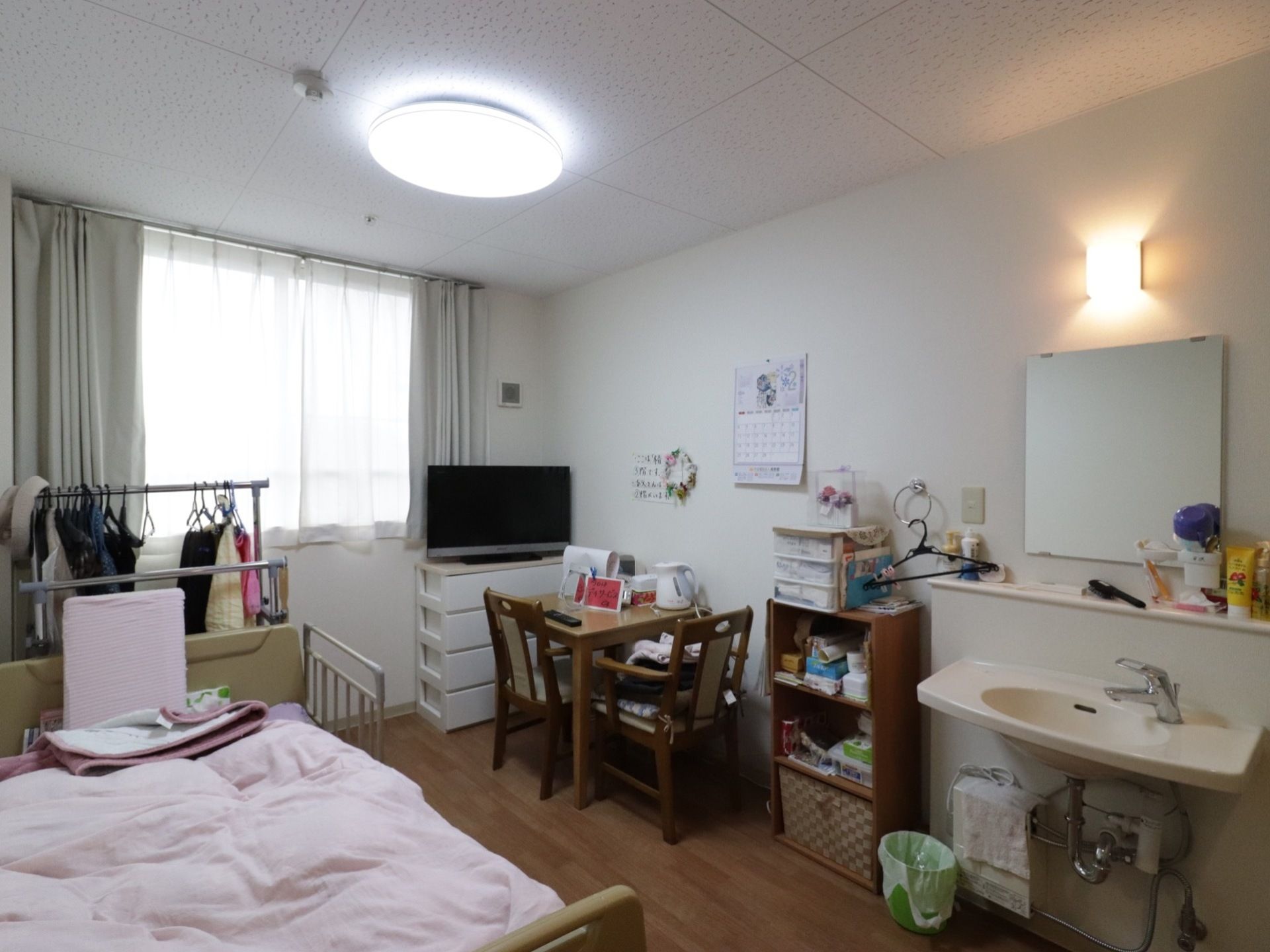 居室にはカーテンがかかった窓がある。テレビやベッド、テーブル、椅子、収納棚、洗面台などが置かれている。