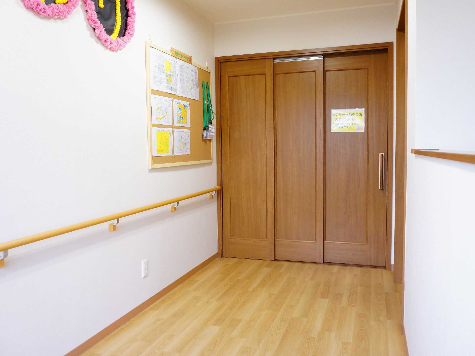 左手の白い壁には手すりが敷設されておりコルクボードなどに掲示物もかけられている。突き当たりには3枚でできたスライド式のドアも設置されている。