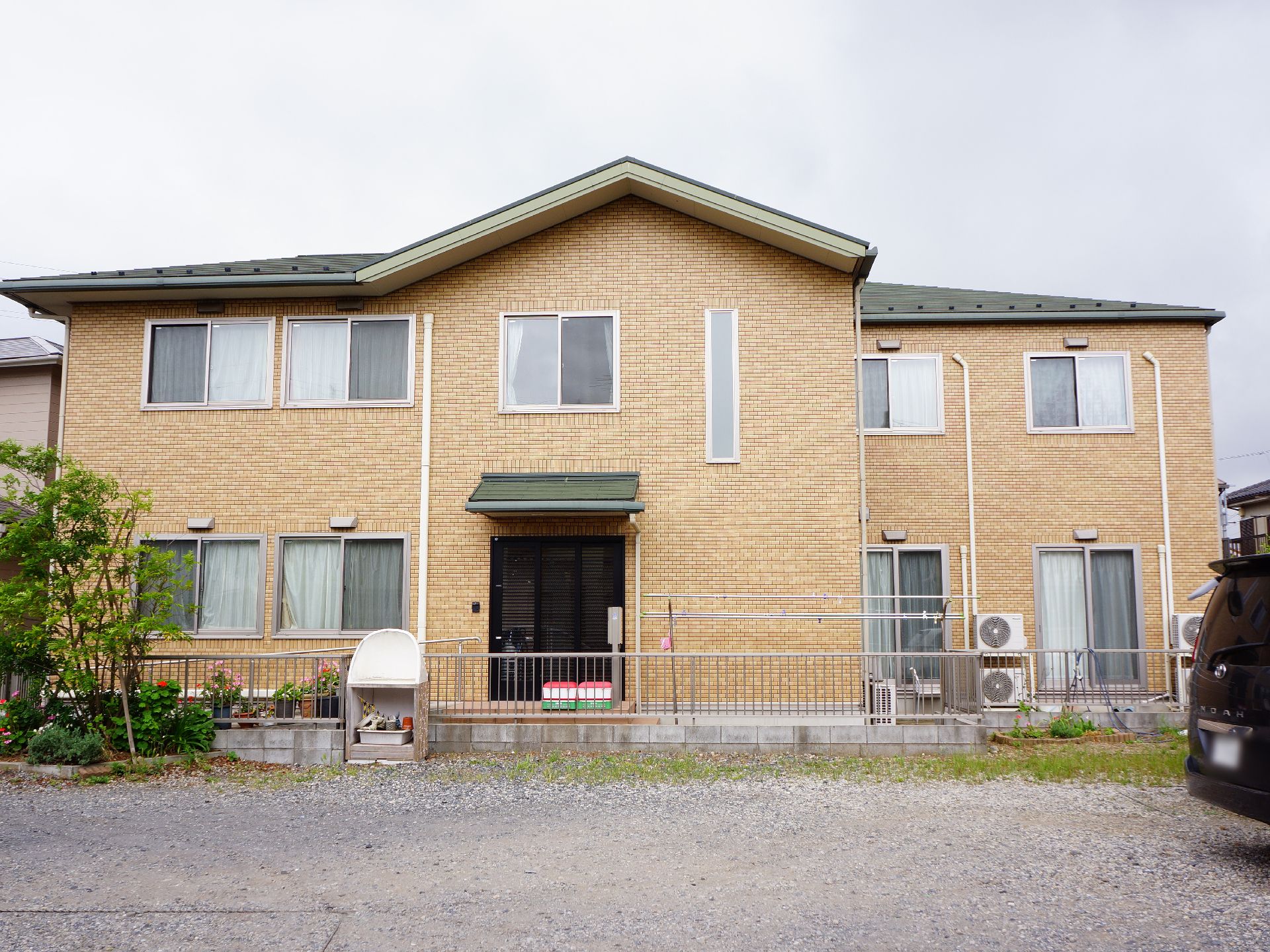 2階建の茶色い外壁の戸建て住宅のような作りをしている。手前には駐車場がありフェンスで隔てられている。