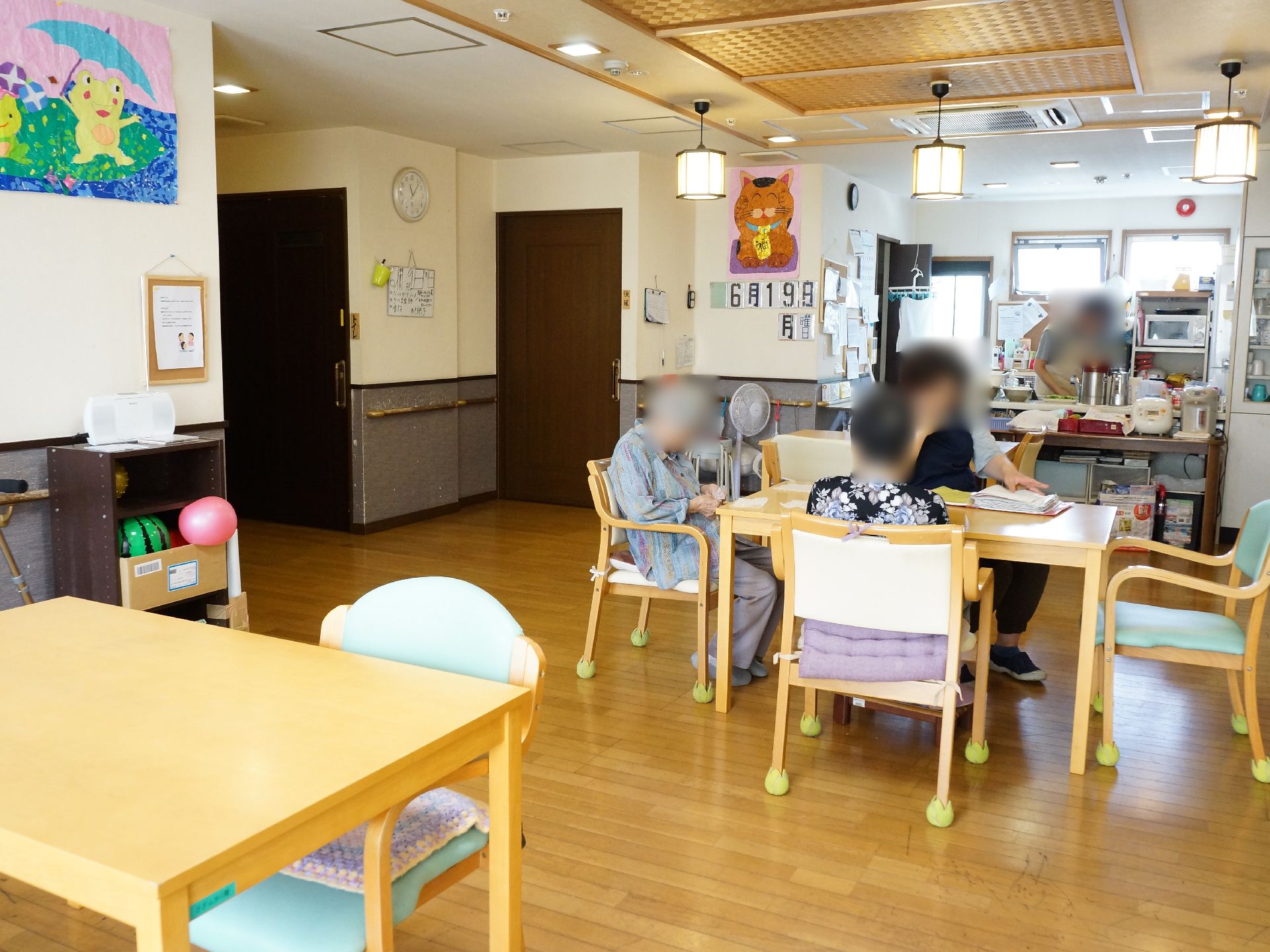 木製のテーブルに入居者が数人集まっている。壁には絵や月日が飾られ天井からはランプシェードが下がっている。
