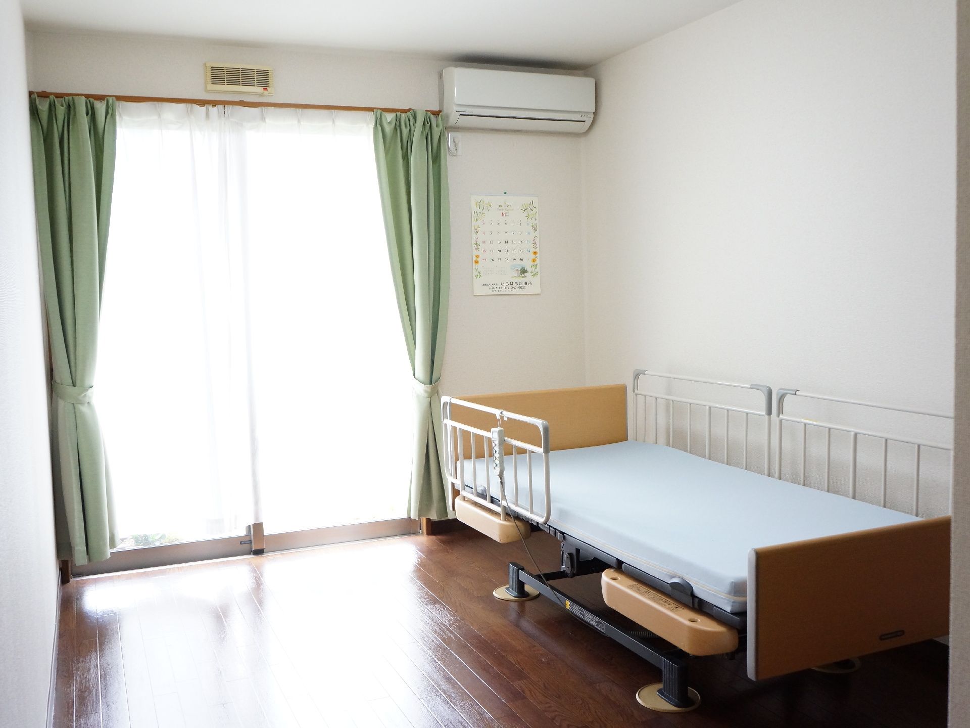 緑のカーテンがかかった部屋に明るい光が差し込んでいる。右上の壁にはエアコンが設置されており、下部には手すり付きのベッドが備え付けられている。