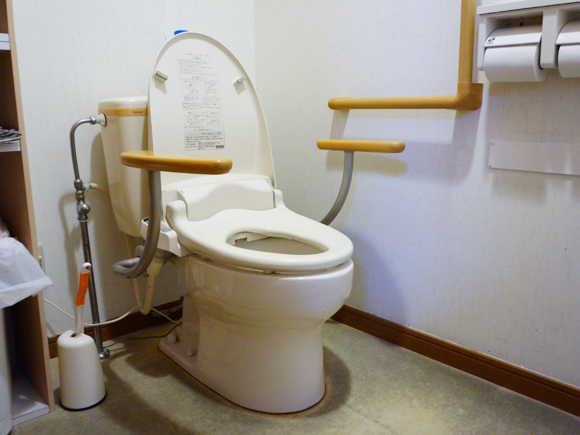 便座の両脇や壁に明るい茶色の介助バーが設置されている。便器の横に棚があり、下の段にゴミ箱が置かれている。