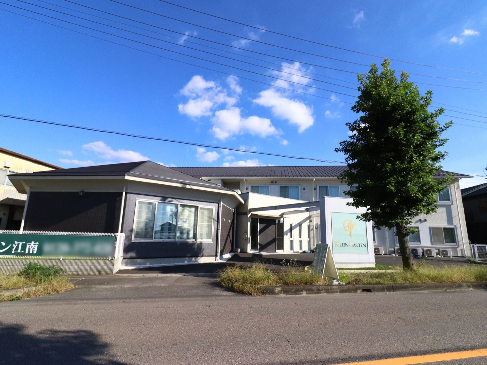 施設手前にある管理棟の建物は黒で、奥の建物は２階建ての白い建物である。施設の前には幹線道路があり、大きな木が一本立っている。