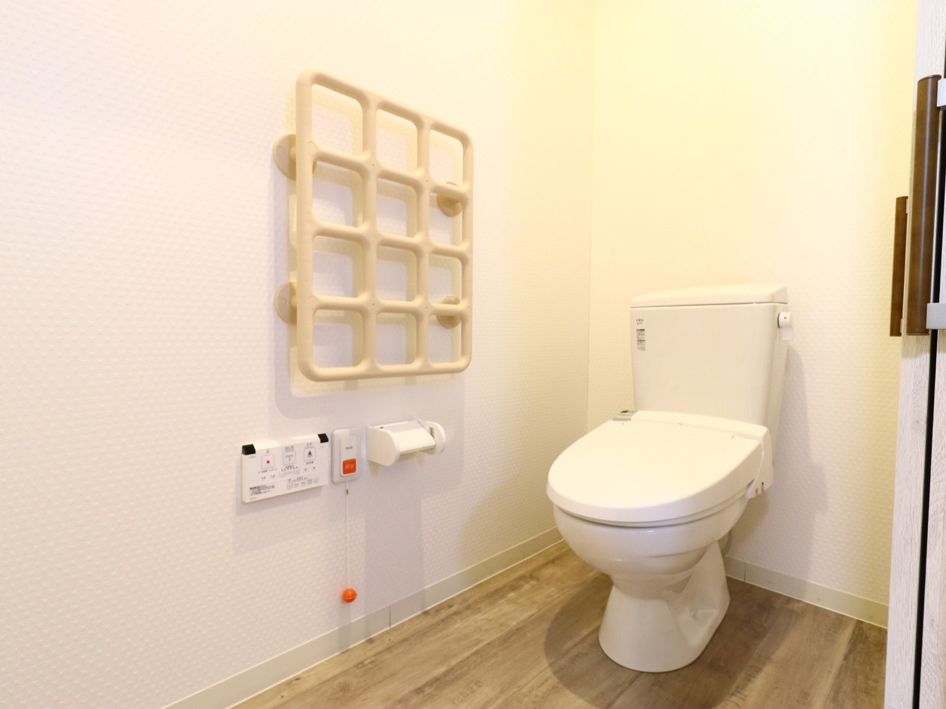 トイレには温水洗浄便座が設置されている。手すりは格子の形をしていて、壁に設置されている。緊急呼び出しボタンもある。