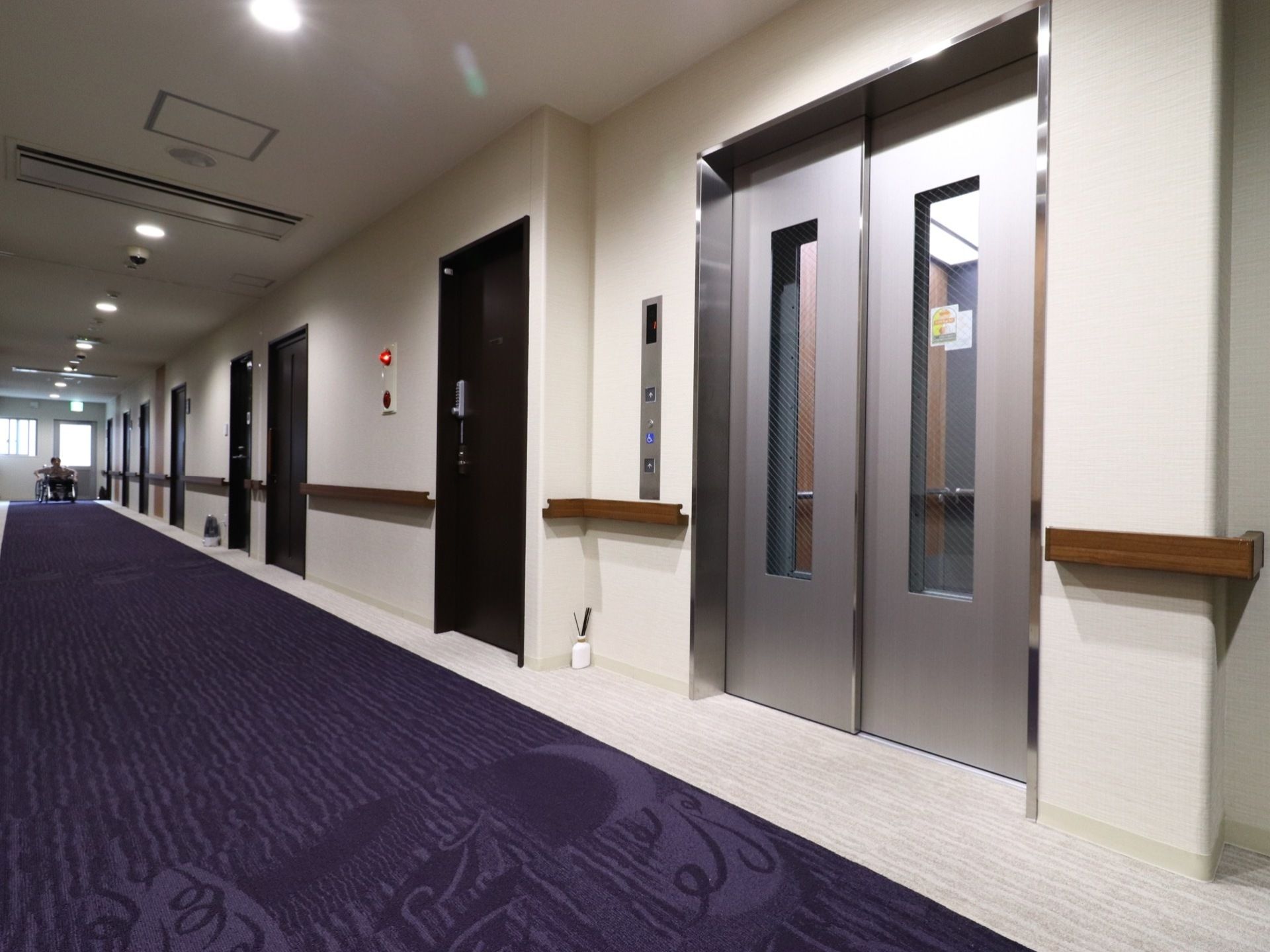 居室のあるフロアーの廊下は長く続いていて、部屋に通じる黒いドアが数多くある。エレベーターには車いす用の押しボタンもある。