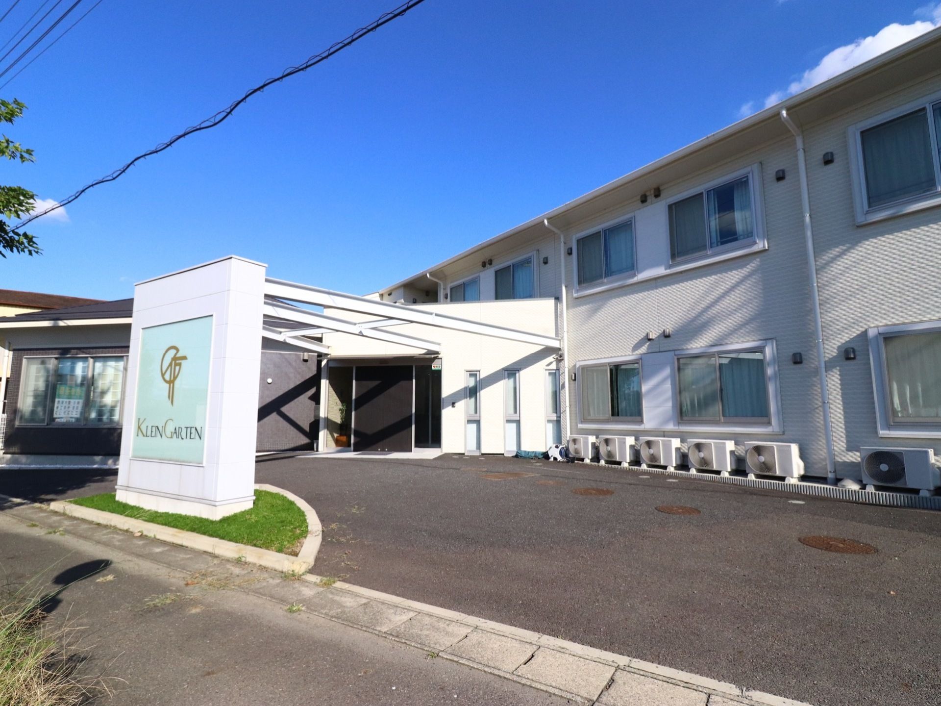 施設は白い２階建ての建物で、１階の入口前には広い駐車スペースがある。通りに面した場所に施設の看板が立っている。