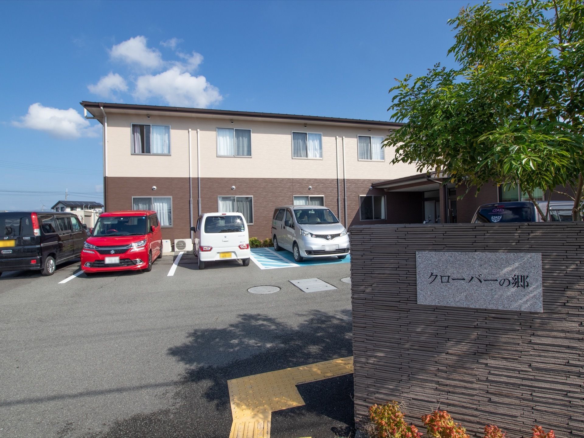 施設は、白色と茶色のツートンカラーの外壁となっている。正面の駐車場には、たくさんの車が停められている。