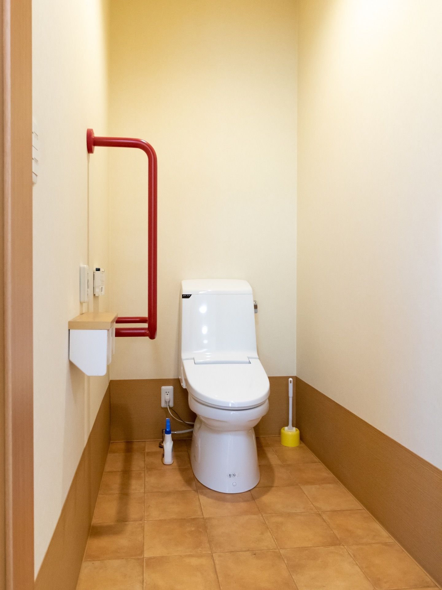 清潔感のある個室トイレで、便器の近くには掃除用具が用意されている。壁には、赤い手すりが取り付けられている。