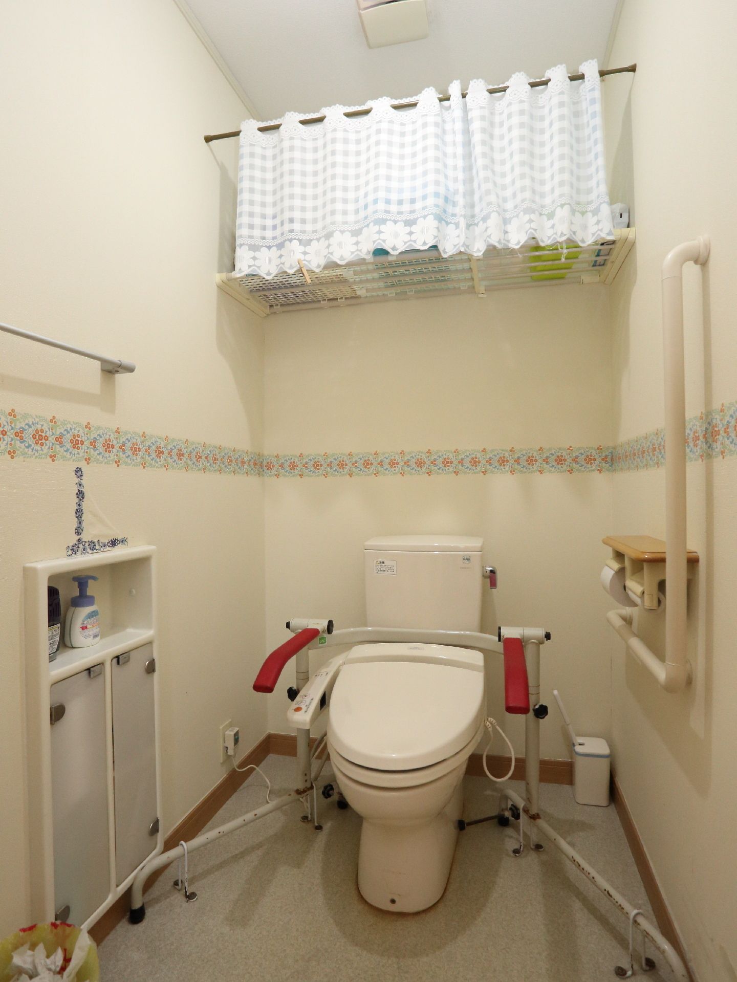 水洗の洋式トイレが置かれている。便座には可動式の手すりがつけられ、ペーパーホルダーの周りにもL字の手すりが設置されている。