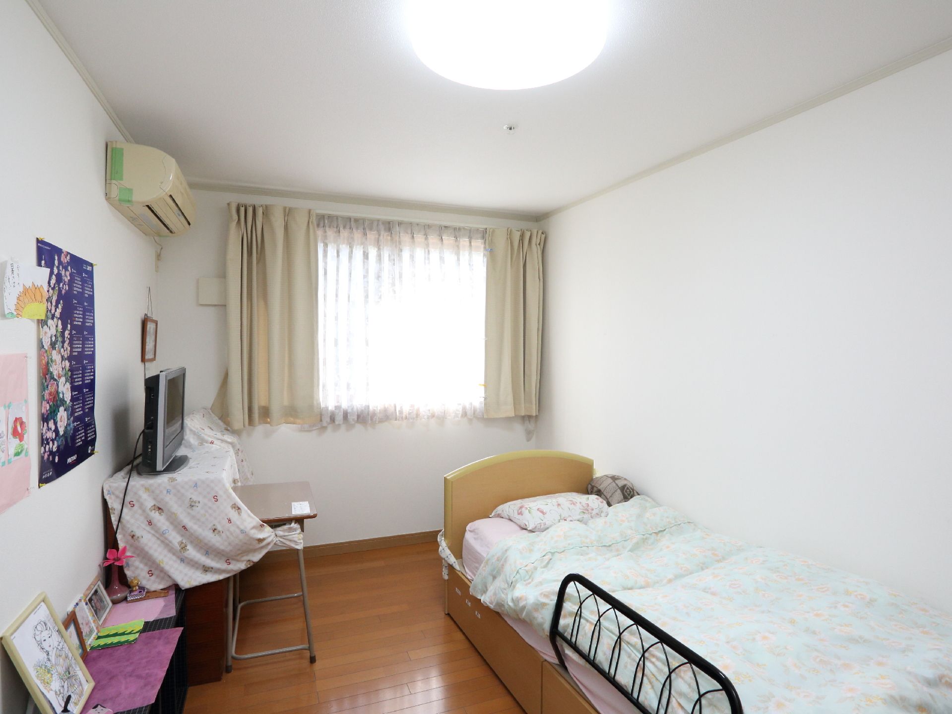 居室の使用例。窓から光の差し込む部屋に、電動ベッドとテレビ、収納台などが置かれている。また、壁や台上は、カレンダーやマットがある。