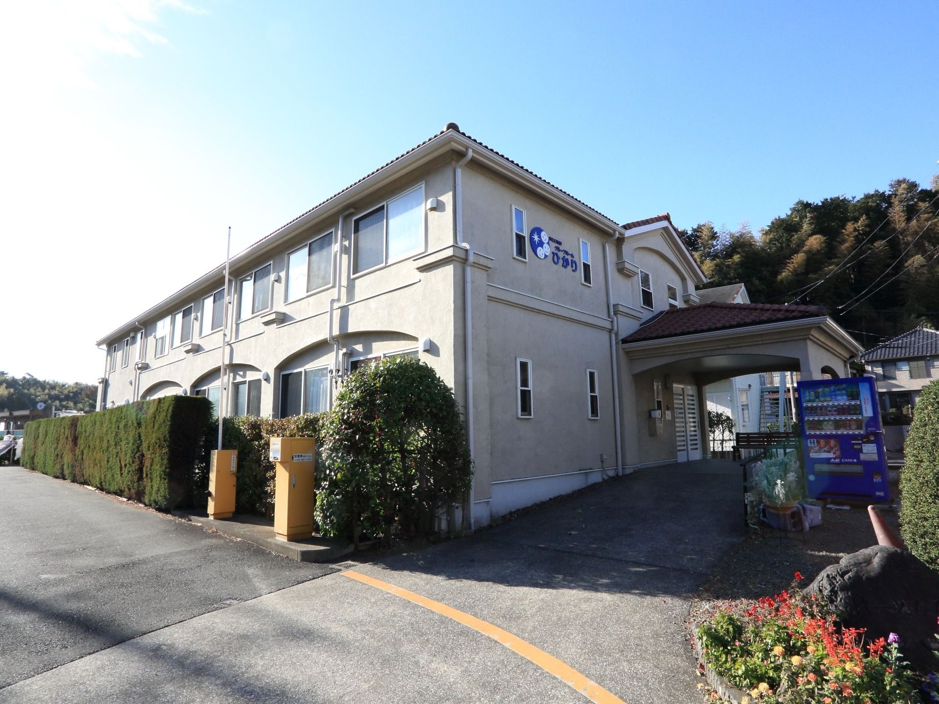 施設の外観の様子。入り口付近には自動販売機が置かれている。施設の周りには、綺麗に整えられた生垣がある。