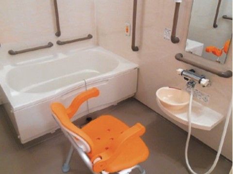 浴槽の横の壁にはナースコールが取り付けられている。シャワーの前にはオレンジ色のバスチェアが置かれている。