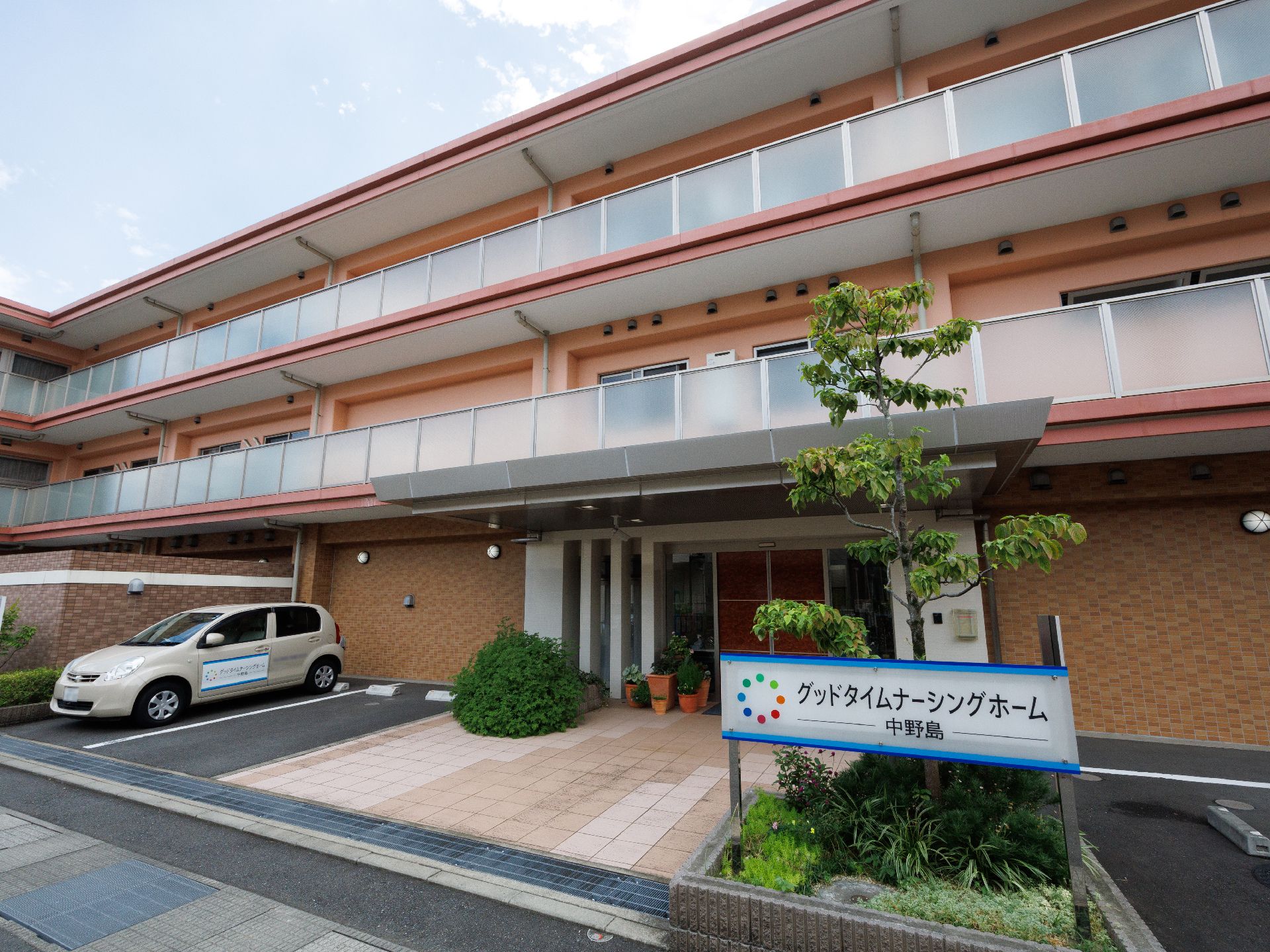 車が止まった駐車場に隣接しているレンガ調でバルコニーが付いた３階建ての建物