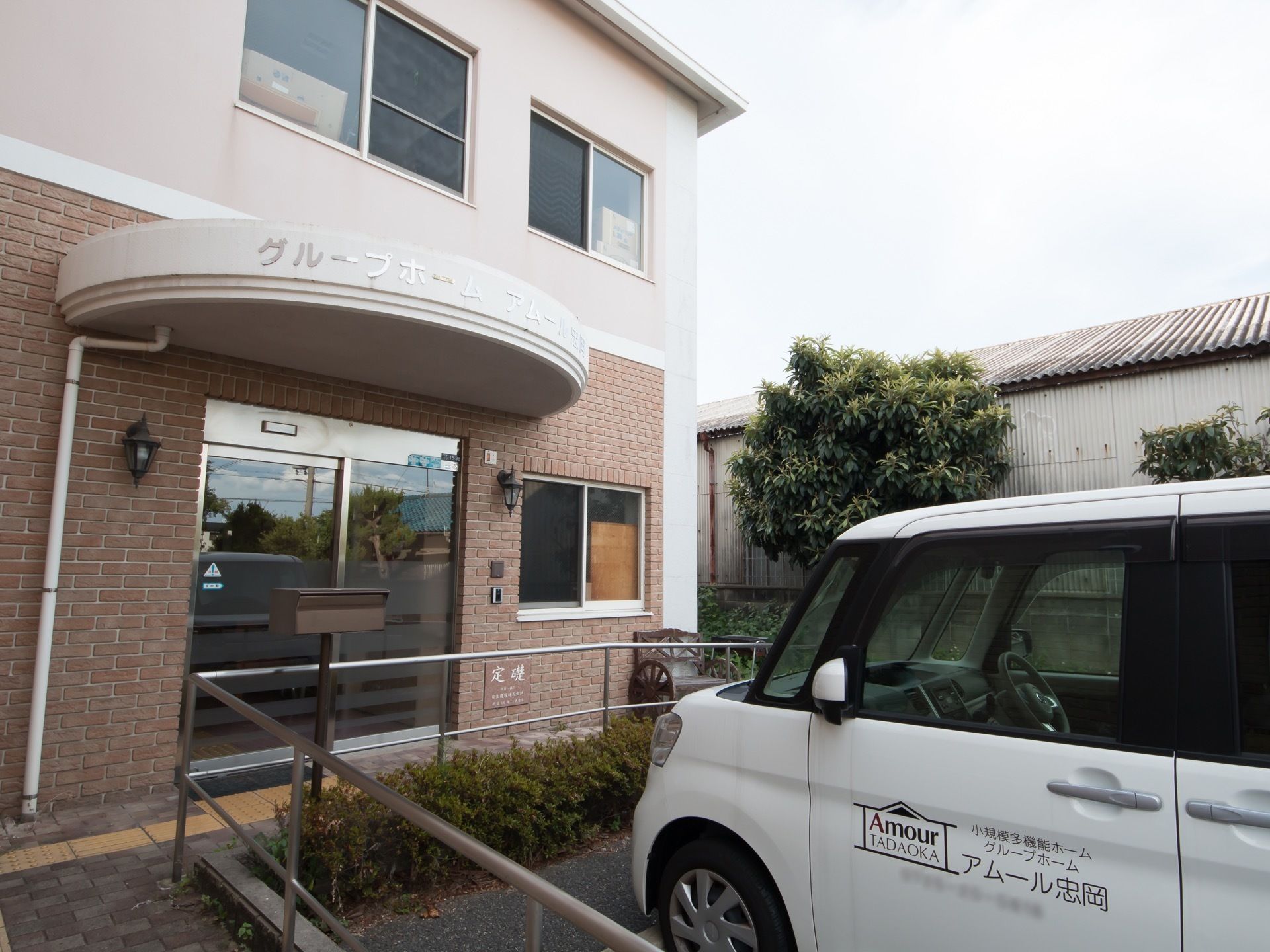 玄関の前には駐車場があり、施設の車が停まっている。植え込みの中に茶色い郵便受けが立っている。駐車場を囲むように手すりが設置されている。