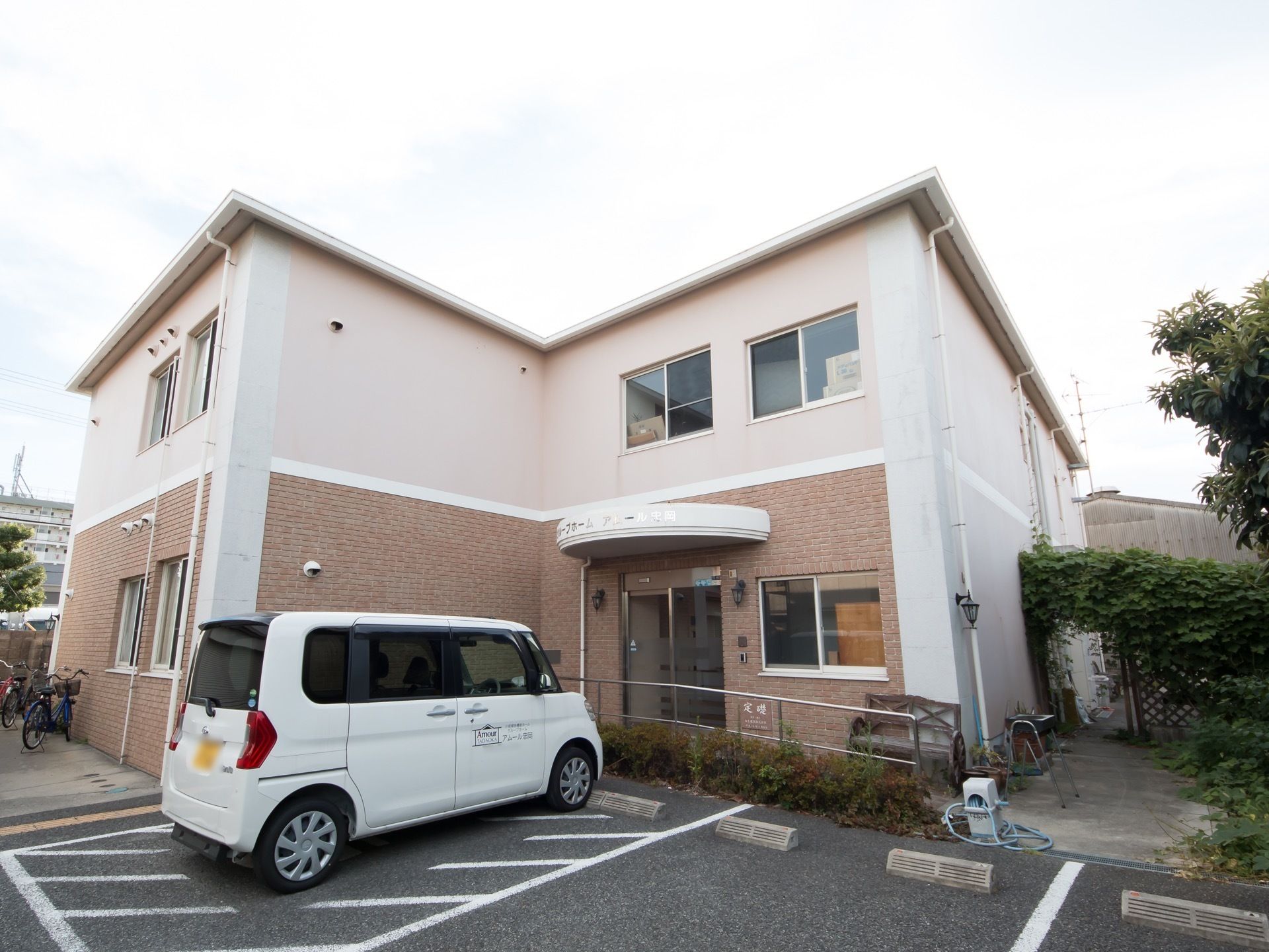 ２階建ての建物で、１階部分はレンガ調の外壁になっている。玄関の前は駐車場になっており、建物の横には自転車が置かれている。