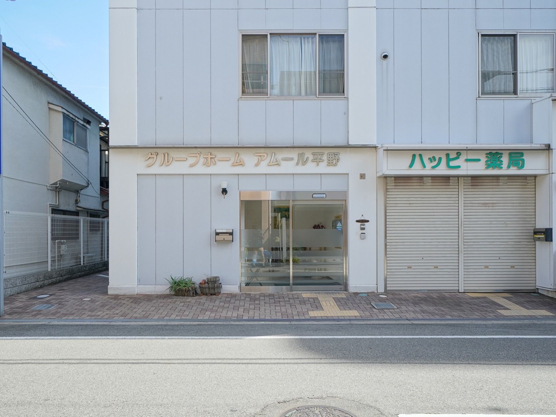 施設の玄関である。大きな自動扉があり、扉の脇には郵便受けがあり、その下に植物の鉢植えが置かれている。隣には薬局がある。