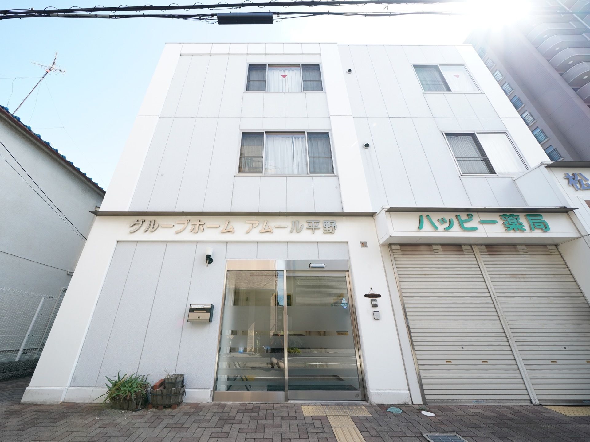 白い３階建てのビルである。施設の玄関は向かって左端である。自動ドアの横に外灯と郵便受けが設置されている。隣には薬局がある。