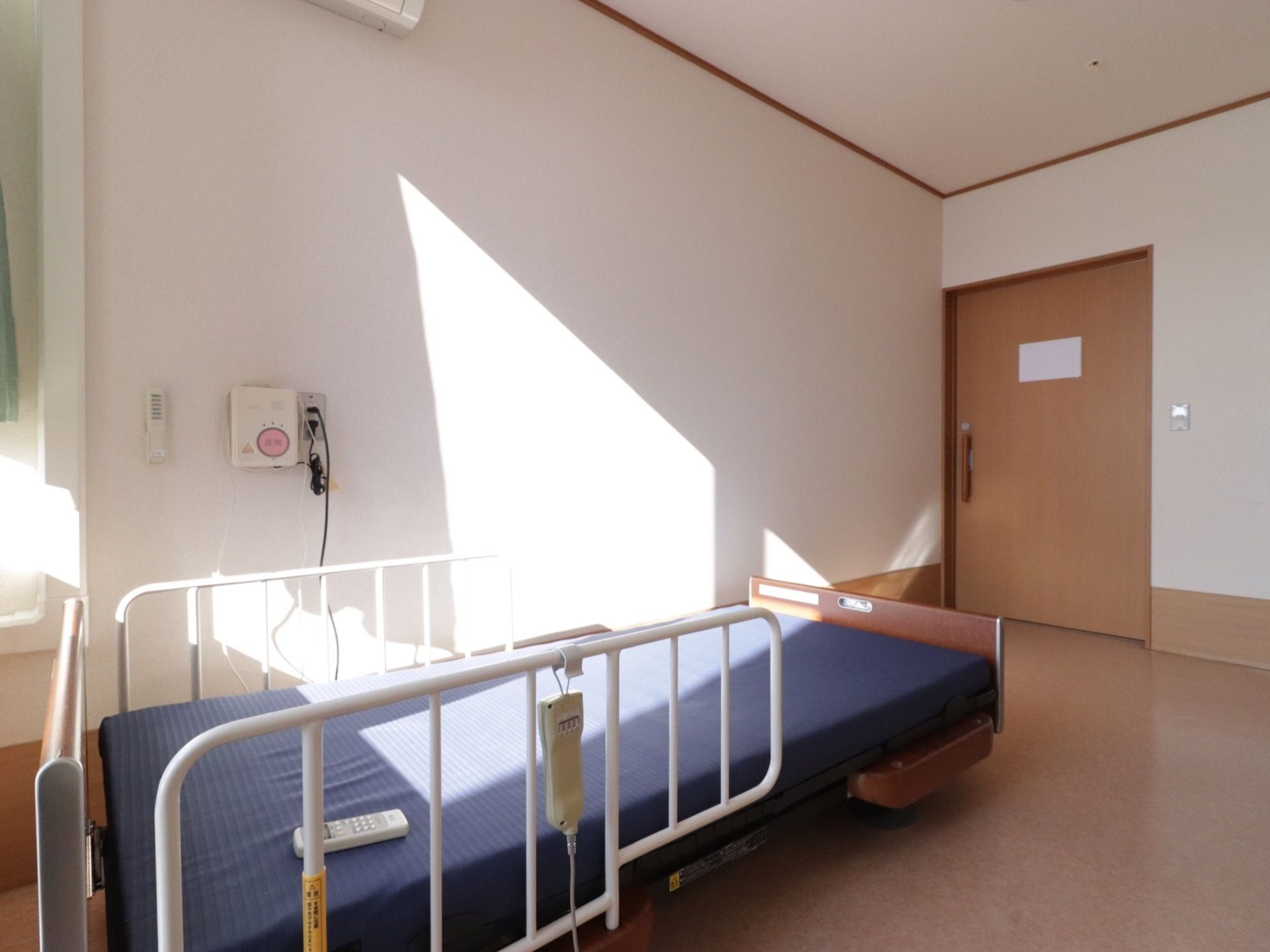 陽射しが入る居室は緊急通報装置が完備されている。マットレスカバーが付けられてた介護用ベッドが設置されており居室の扉は木製の引き戸である。