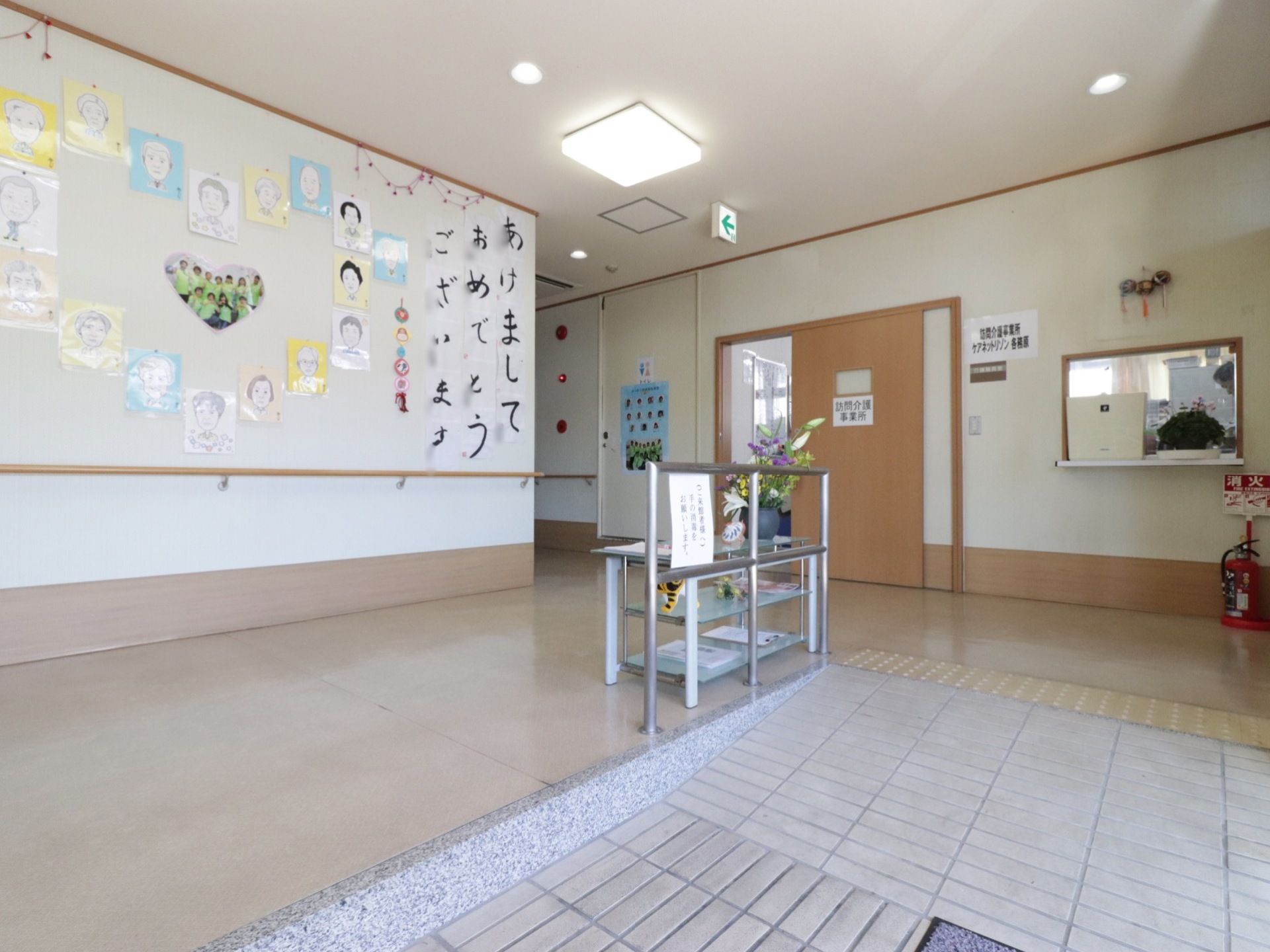 玄関は移動に安全なスロープが設置されている。壁には入居者様全員の似顔絵とスタッフの全員の写真が貼られている。