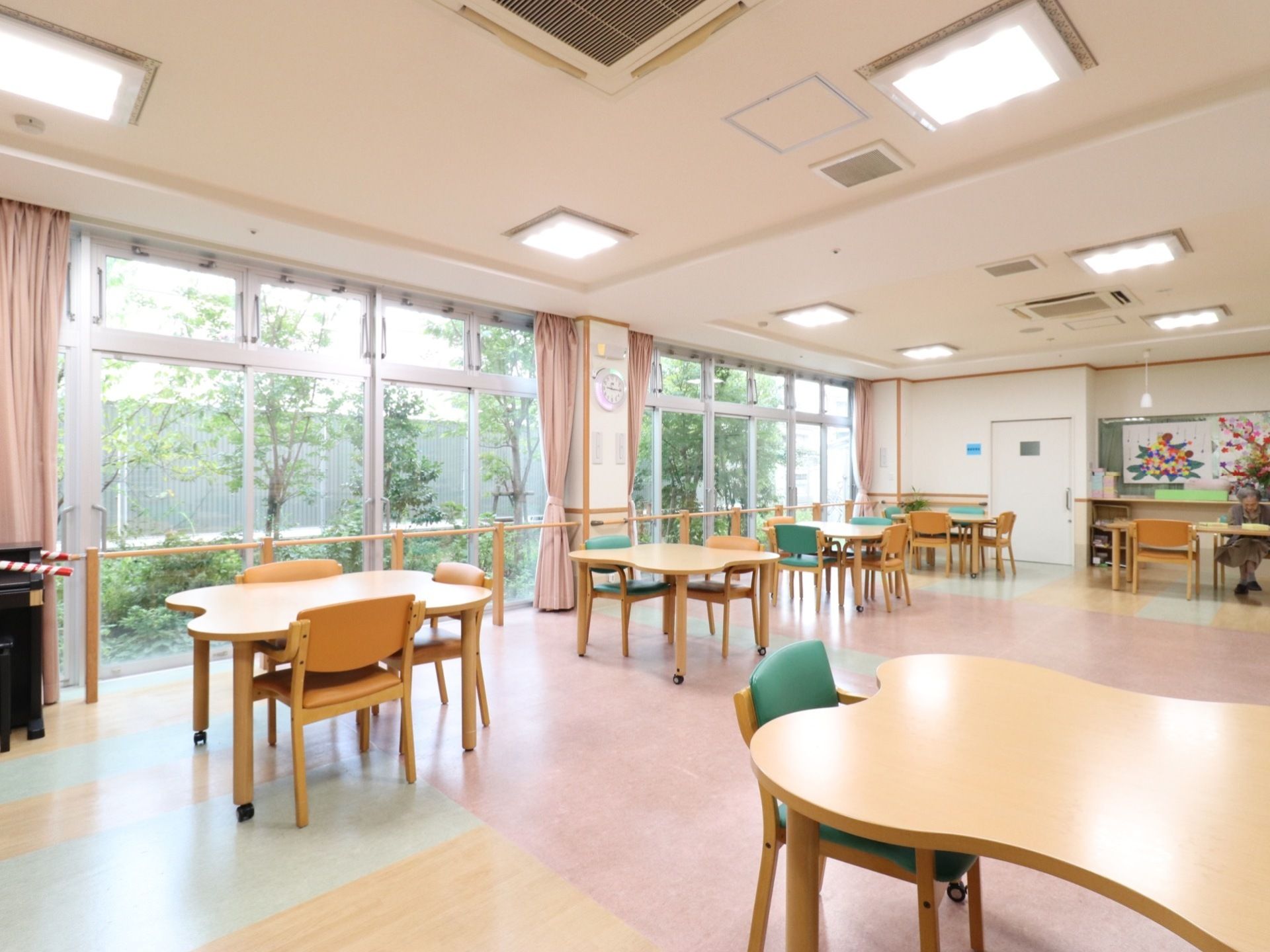 大きな窓から光が入り明るい空間。緑も見えて寛いで過ごせる。窓際に平行棒があり、歩行訓練もできる場所になっている。