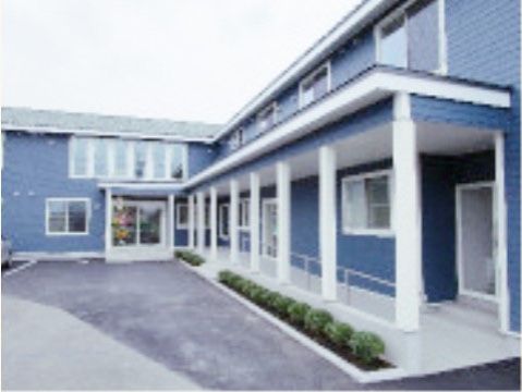 青と白に塗られたお洒落な２階建ての建物。玄関前には手すり付きのスロープがあり、脇には植物が植えられている。