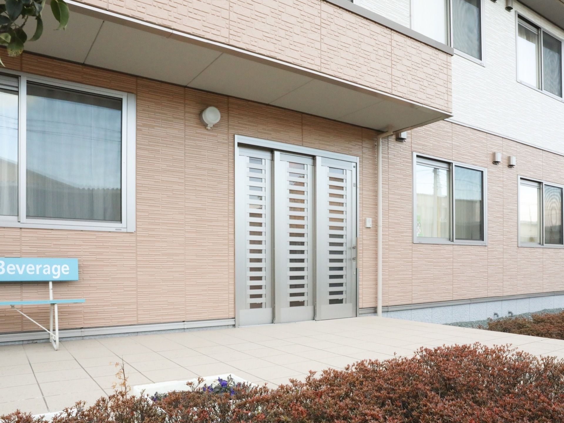 建物の外壁は、ベージュ色でまとめられた落ち着いたデザインである。金属製の自動ドアの左上には、丸い形の照明器具がある。