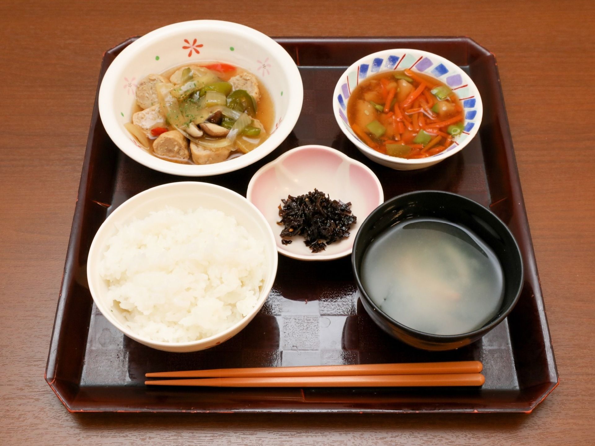 四角いトレーの上に、ご飯、味噌汁、野菜とつみれのスープ煮込み、人参やインゲンの入ったあんかけ料理などが並んでいる。