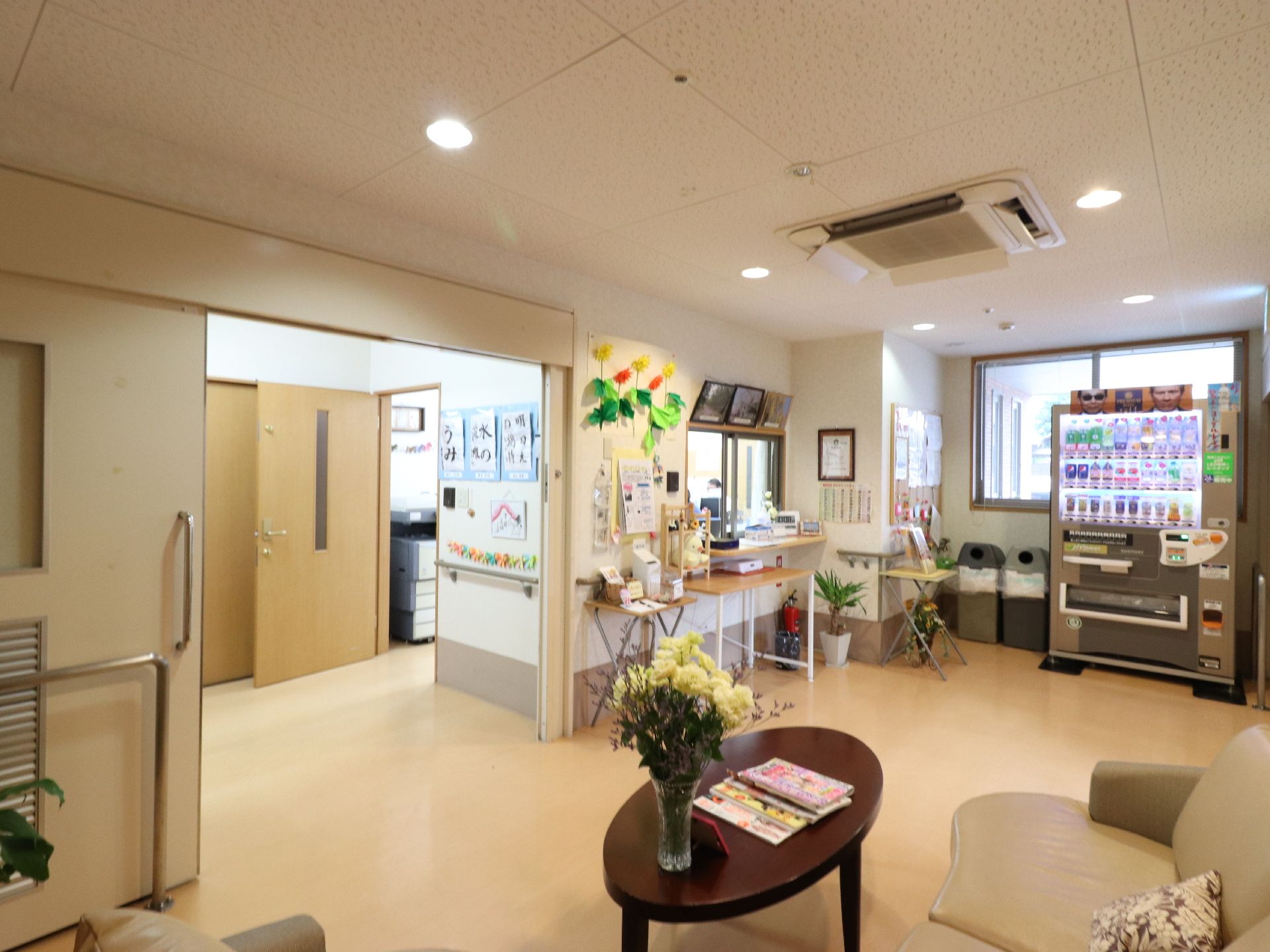 受付左側には、出入り口がある。壁には入居者の習字の作品や折り紙などが掲示されている。天井にはエアコンが設置されている。
