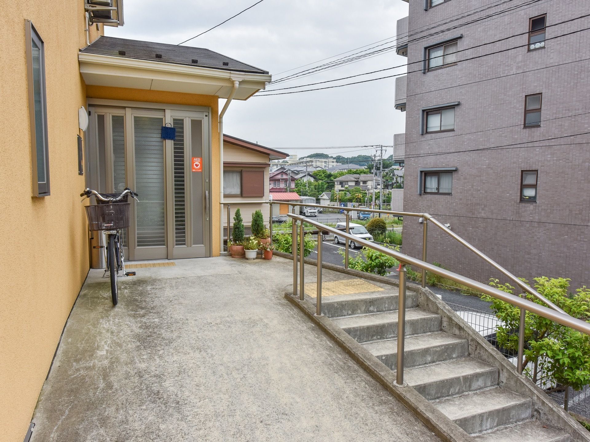 玄関は一般住宅のような雰囲気である。玄関横に自転車と鉢植えの植物が置かれている。階段と手すり、スロープが設置されている。