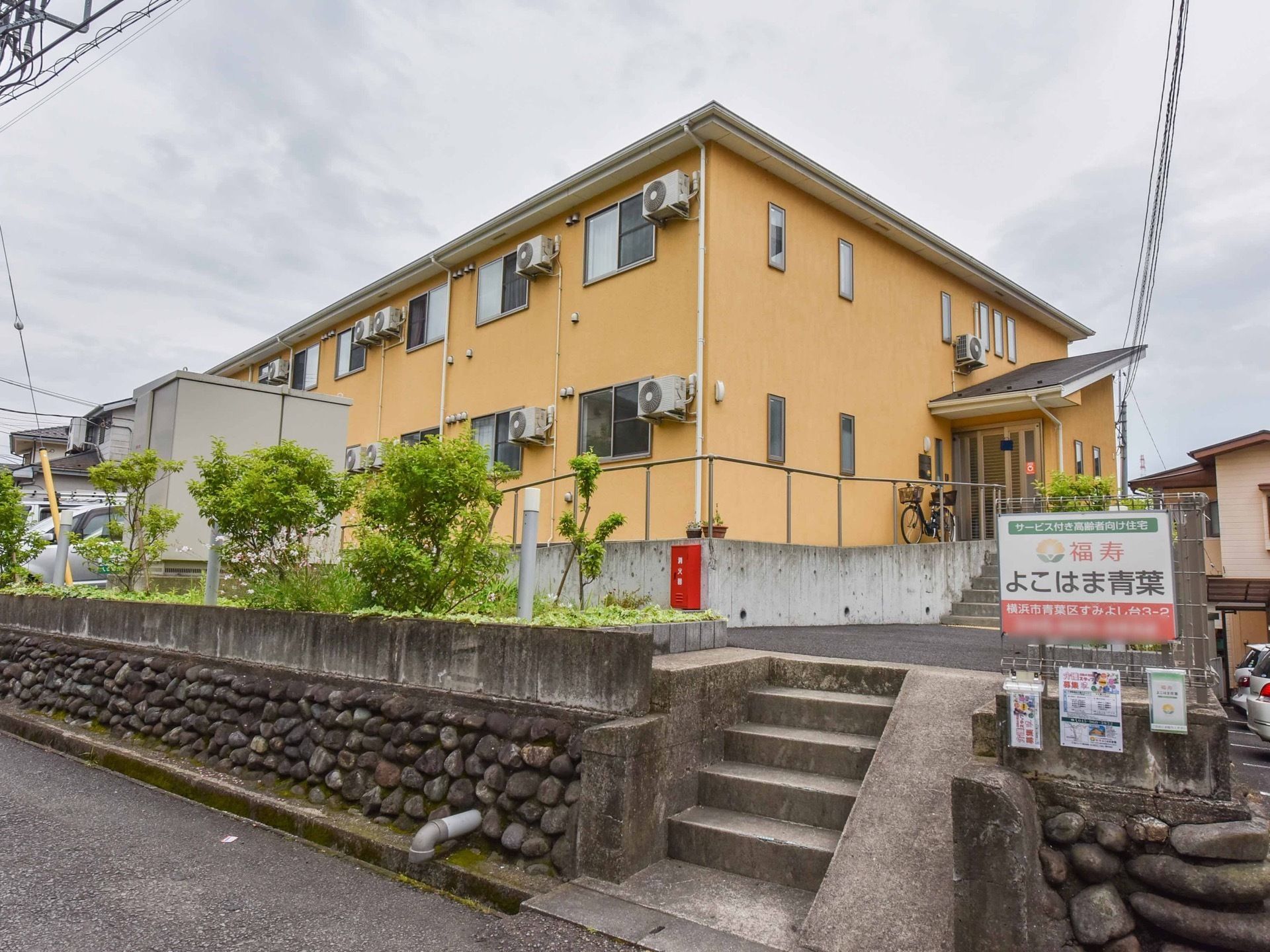 外壁は明るいオレンジ色で２階建ての建物である。下の道路から階段が続いていて、階段の横に施設名の看板が設置されている。