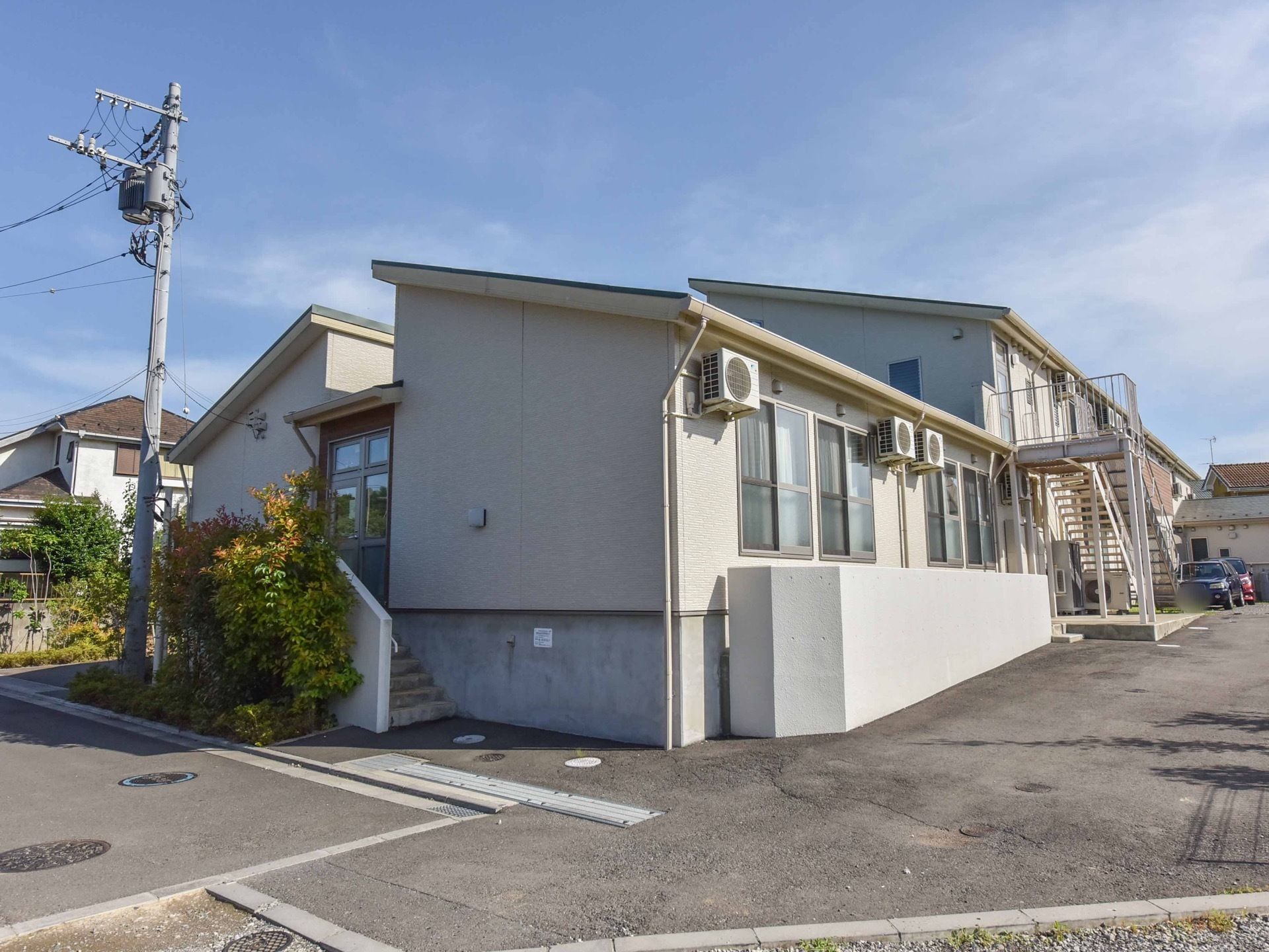町並みに溶け込むような、ベージュを基調とした外壁の木造２階建ての建物。裏手にアスファルトの広いスペースが設けられている。
