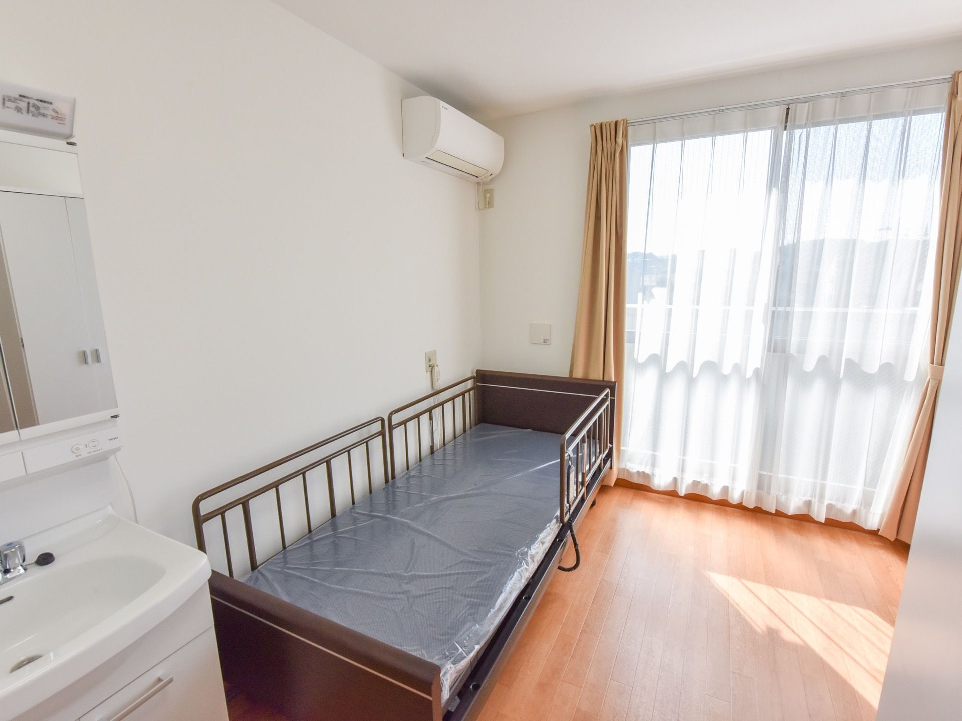 木目調のフローリングの部屋に柵付きの茶色のベッドとエアコン、化粧鏡付きの洗面台が設置されている。窓にはレース・遮光のカーテンがある。